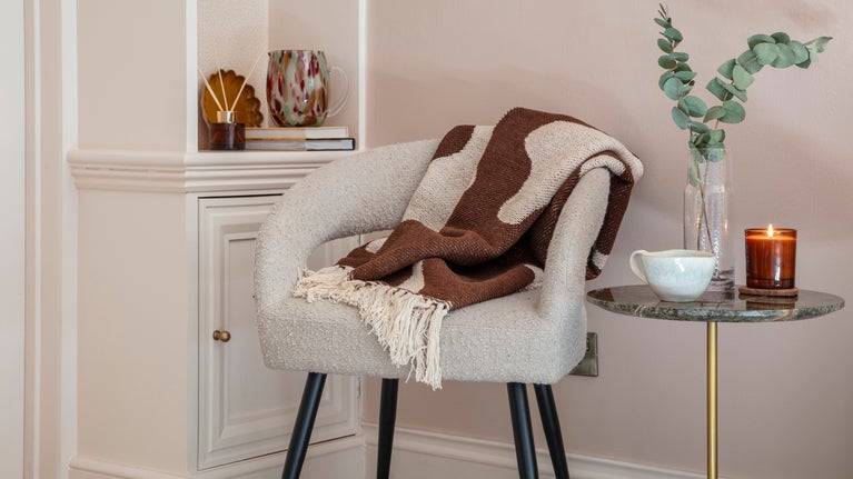 A cream and brown throw displayed on a cream boucle chair. A lit candle and a cup of tea are placed on a side table.